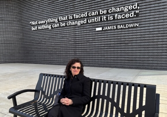 Mindy Tarlow sitting on a bench in front of a wall featuring a quote by James Baldwin: “Not everything that is faced can be changed, but nothing can be changed until it is faced.”